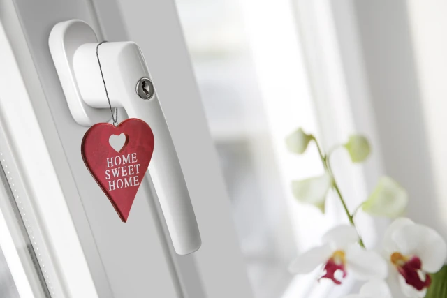 Detailaufnahme eines WIRUS Fensters mit Fokus auf den abschließbaren Griff als Einbruchschutz; daran hängt ein rotes Herz mit der Aufschrift „Home Sweet Home“, im Hintergrund eine helle Wohnszene mit Blumen.